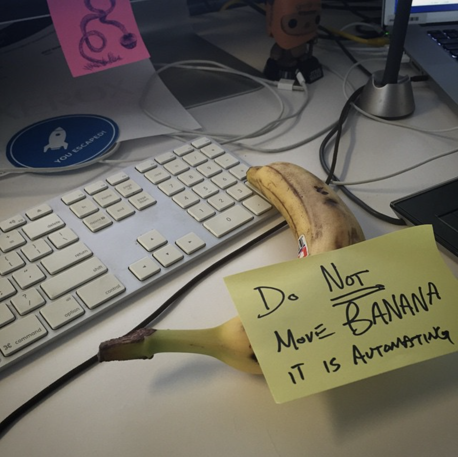 Banana on desk with yellow sticky note reading 'Do NOT Move BANANA IT IS AUTOMATING' next to keyboard